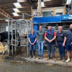 Quatre éleveurs et une éleveuse devant la stalle d'un robot de traite d'où sort une vache de race Holstein